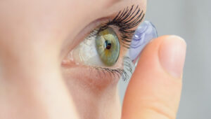 Zoom nas mãos de pessoa caucasiana aproximando uma lente de contato de grau dos olhos com o dedo indicador.