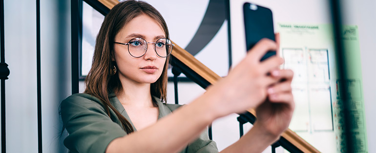 Imagem de mulher caucasiana usando ócuos de grau redondo enquanto segura smartphone tirando selfie.
