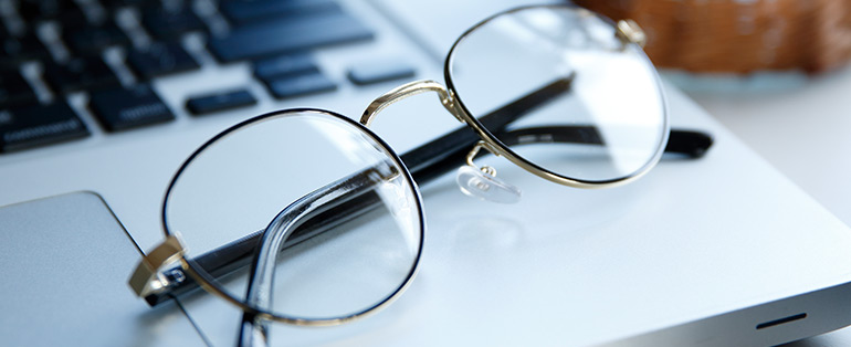 Imagem de óculos arredondado apoiado na base de um laptop representando as lentes fotossensíveis.