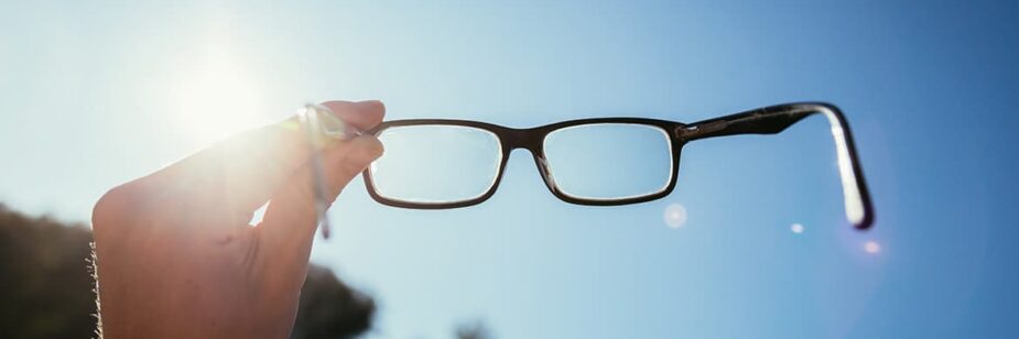 Imagem de mão segurando um par de óculos na direção do sol com o céu azul no fundo