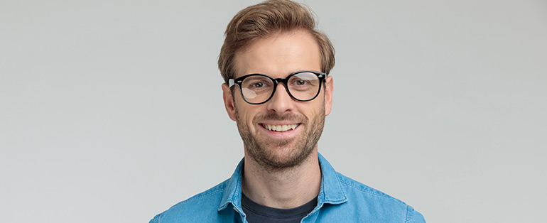 Imagem de homem caucasiano e loiro, com a barba por fazer e sorrindo. Ele usa óculos de grau redondo com armação preta.