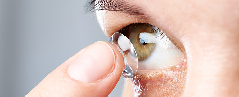 Imagem com zoom no olho de uma pessoa caucasiana colocando lentes de contato em um dos olhos com a ponta do dedo indicador. 