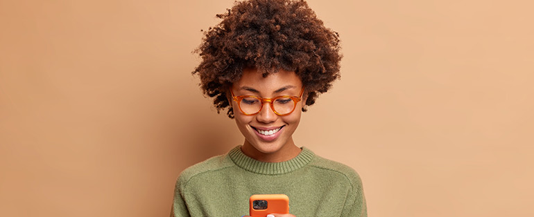 Mulher negra usando óculos de grau com armação na cor laranja posa em frente a fundo bege enquanto olha celular 