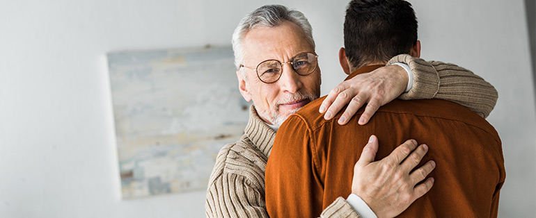 Homem idoso caucasiano com cabelos brancos usando óculos de grau abraça outro homem de costas.