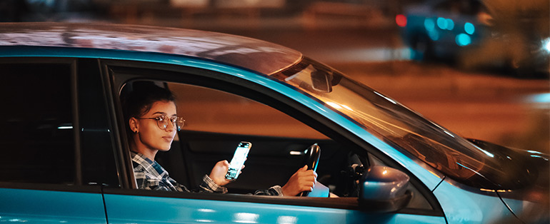 Imagem de mulher caucasiana dentro de carro usando óculos com uma das mãos no volante e outra no celular. 
