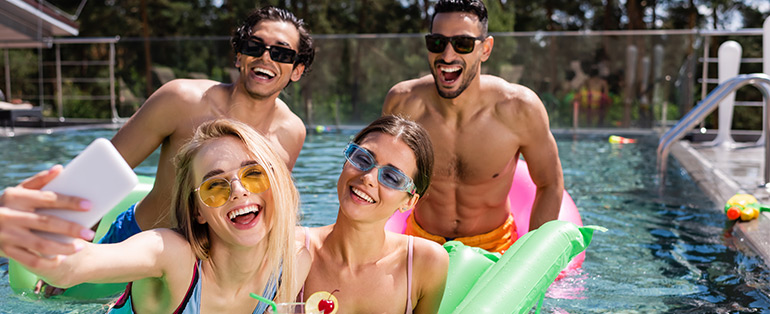 Imagem de quatro amigos caucasianos, duas mulheres e dois homens, tirando selfies dentro de piscina enquanto usam óculos de sol. 