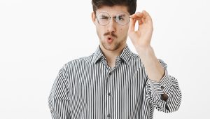 Homem caucasiano com camisa listrada segura óculos de grau redondos em frente ao rosto verificando se existem riscos ou arranhões.