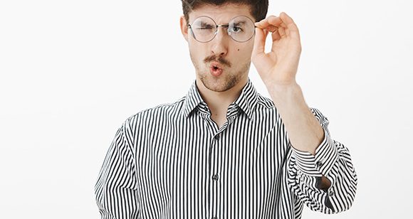 Homem caucasiano com camisa listrada segura óculos de grau redondos em frente ao rosto verificando se existem riscos ou arranhões.