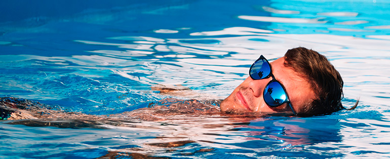 Homem caucasiano com o corpo dentro de piscina enquanto usa óculos de sol com lentes espelhadas