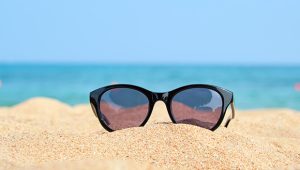 Óculos de sol no formato gatinho apoiado na areia de praia com o oceano ao fundo.