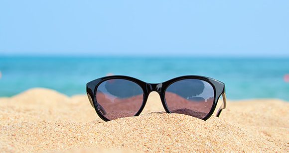 Óculos de sol no formato gatinho apoiado na areia de praia com o oceano ao fundo.