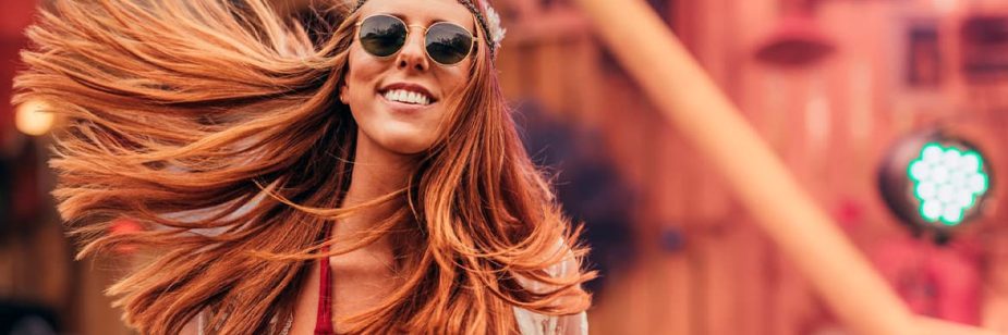 Vemos uma mulher sorridente com oculos de sol redondo no rosto com os cabelos ruivos voando e um arco pendurado na cabeça