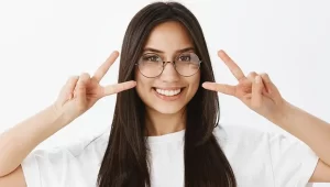 Uma mulher sorridente fazendo o sinal de dois com as duas mãos usando um óculos de grau redondo