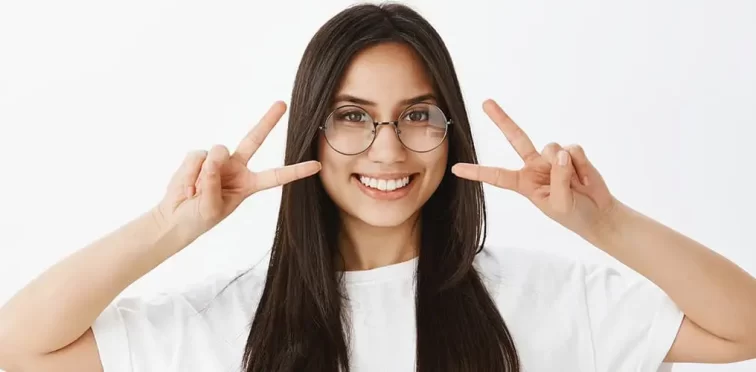 Uma mulher sorridente fazendo o sinal de dois com as duas mãos usando um óculos de grau redondo